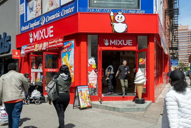A Mixue Ice Cream & Tea shop occupies a bright red corner storefront in Downtown Brooklyn. Large white lettering and the chain's mascot cover the awnings, while pedestrians enter beneath colorful drink posters. Comparing ice cream shop locations by season, foot traffic, and the one question that decides it